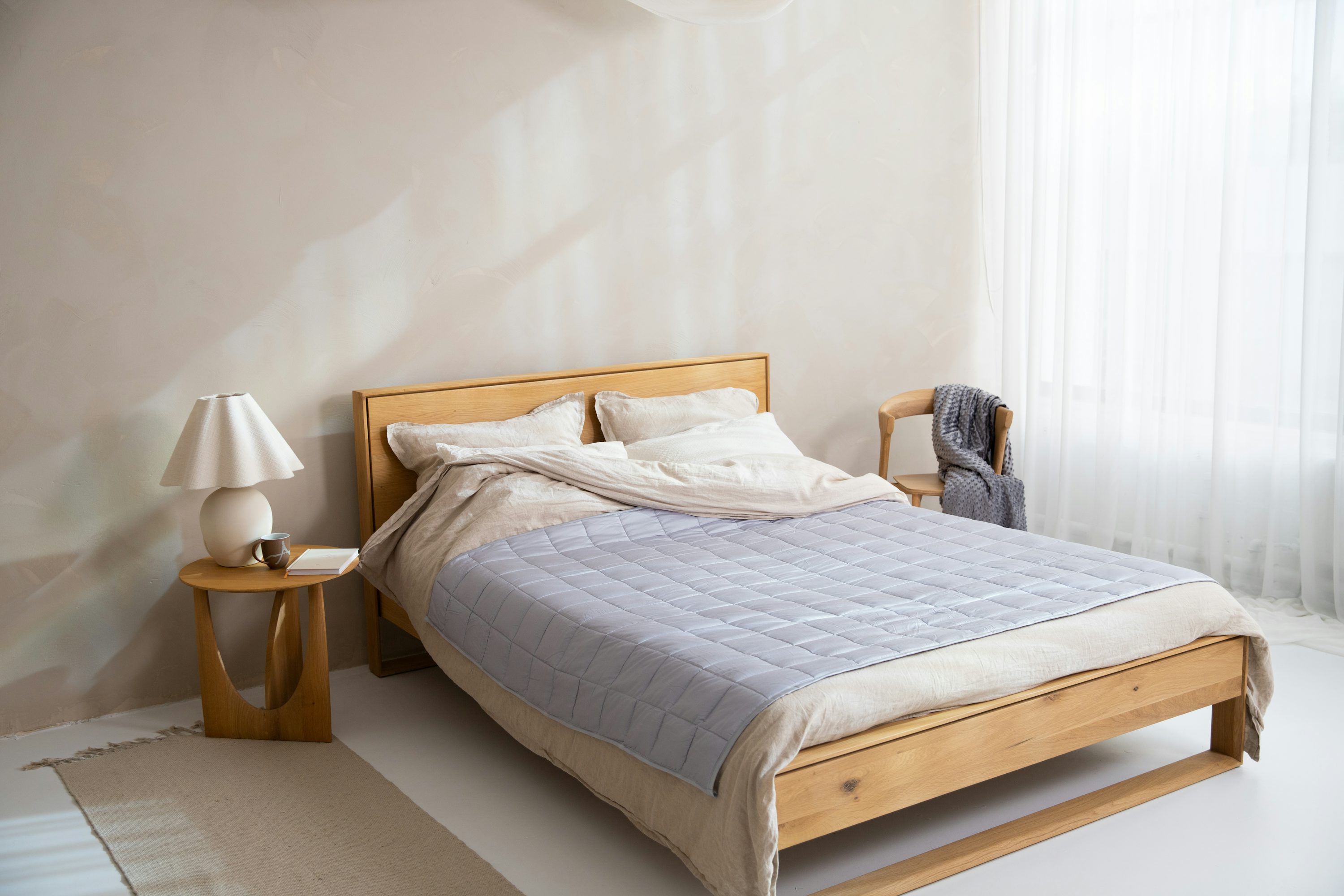 Lit en bois avec couvre-lit gris clair et linge blanc, ambiance calme et lumineuse, le confort apporté par le linge de lit.